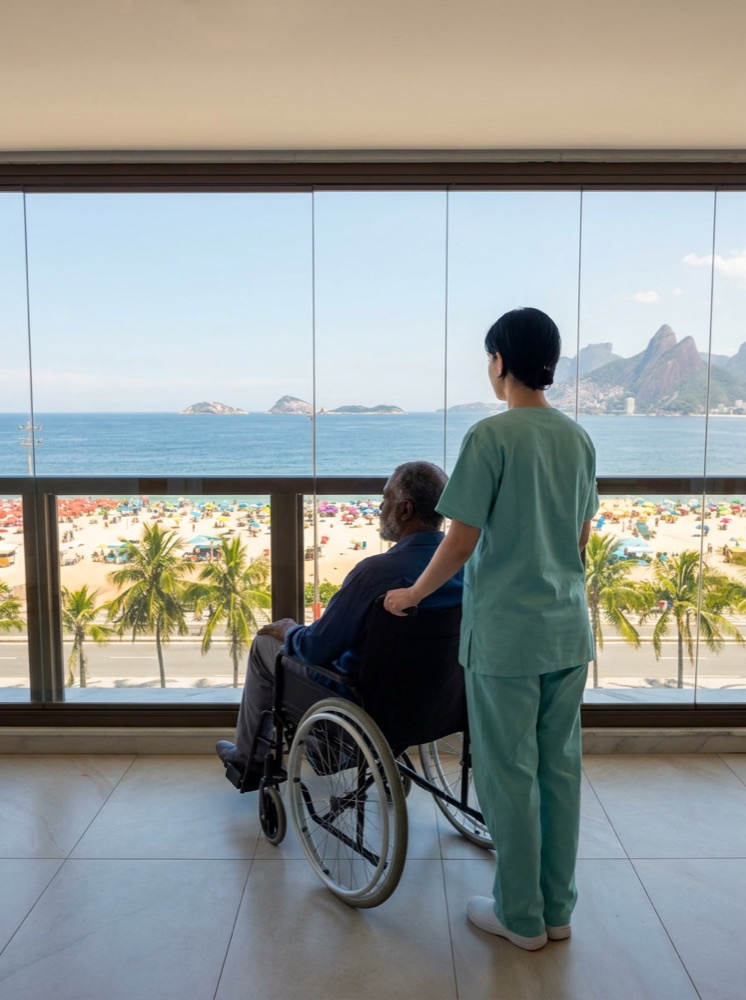 Cuidadora da Santer Saúde levando idosa em cadeira de rodas a um terraço com vista para o Rio de Janeiro